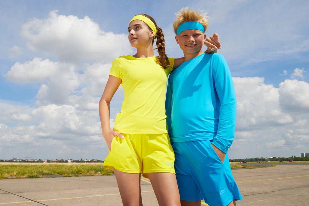 Mädchen mit gelber Sportkindkleidung bestehend aus Capsleeve Shirt, Shorts und Stirnband und Junke mit türkiser Sportkleidung bestehend aus Longleeve, Shorts und Stirnband